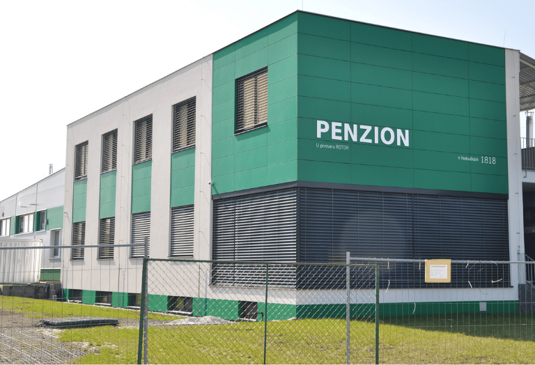 Pohled na budovu penzionu s moderní fasádou v kombinaci bílé a sytě zelené barvy fasádních panelů. Budova má velká prosklená okna s venkovními žaluziemi a je obklopena zelení za drátěným plotem.
