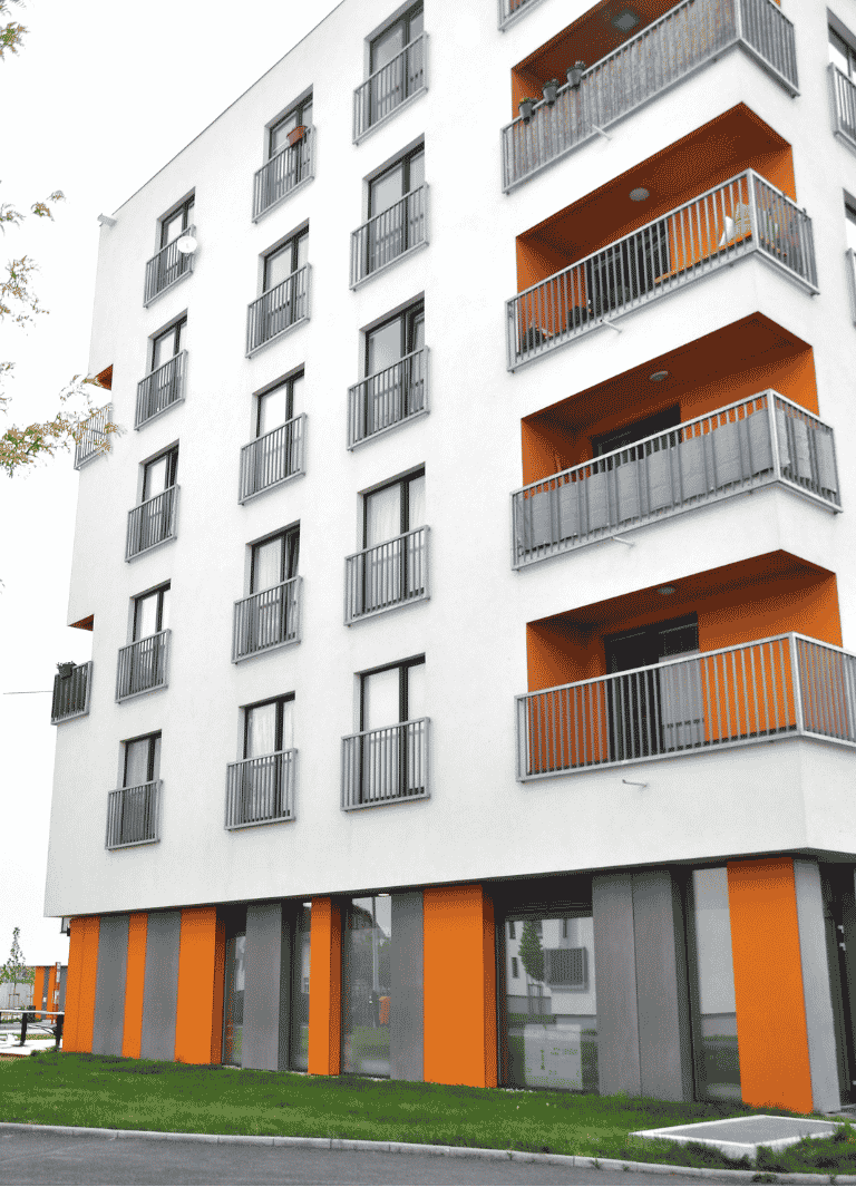 Detailní pohled na moderní bytový dům s čistě bílou fasádou, kterou doplňují hravé akcenty sytě oranžových fasádních panelů v parteru a na balkonech.